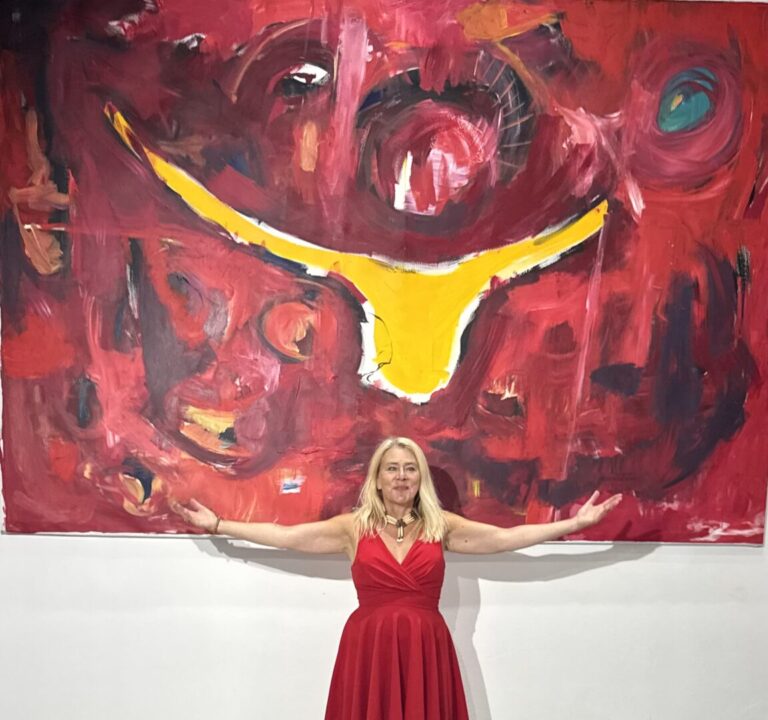 Susanne Rikus in a red dress stands with open arms in front of a large abstract red painting with yellow and dark elements; a wooden and metal sculptural installation lies in the foreground.